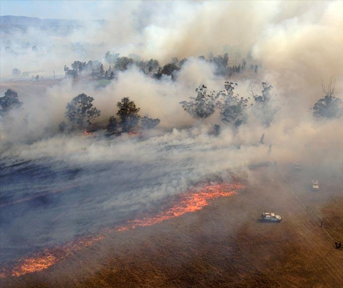 Aerial view of a wildfire with visible flames and thick smoke, spreading across a landscape with trees and a road, where two vehicles are present near the fire line.