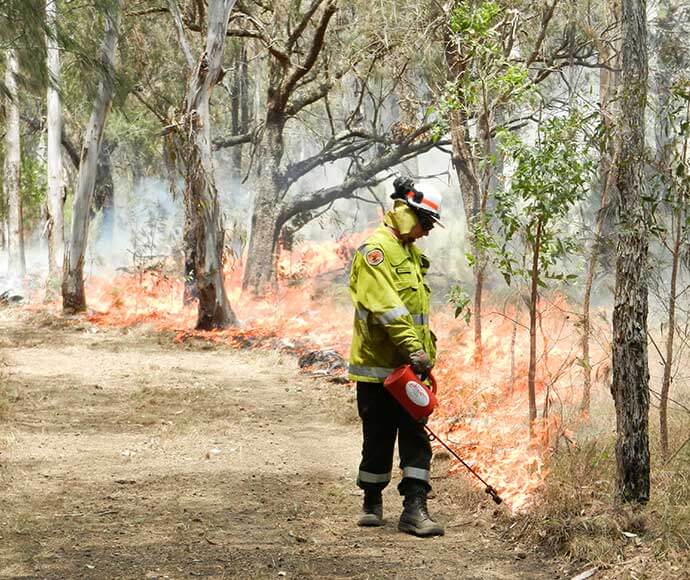 Rural fire fighter wearing protective clothing and helmet, walking down a track and burning the grass as he goes.