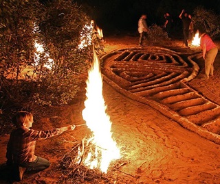 A person is kneeling on the ground, tending to a large fire that outlines an intricate labyrinth pattern. The fire’s glow illuminates the paths of the labyrinth, creating a warm and possibly ceremonial atmosphere. In the background, other individuals are also attending to different parts of the fire or observing. The setting appears to be outdoors at night, and the labyrinth fire adds an intriguing element to the scene.