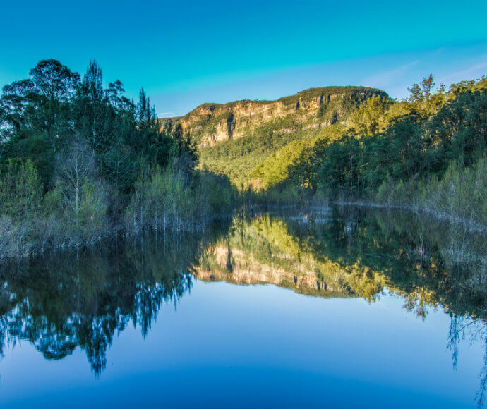 Water with trees lining either side and a mountain in the background. The water is glassy and has a perfect reflection of the trees and mountains.