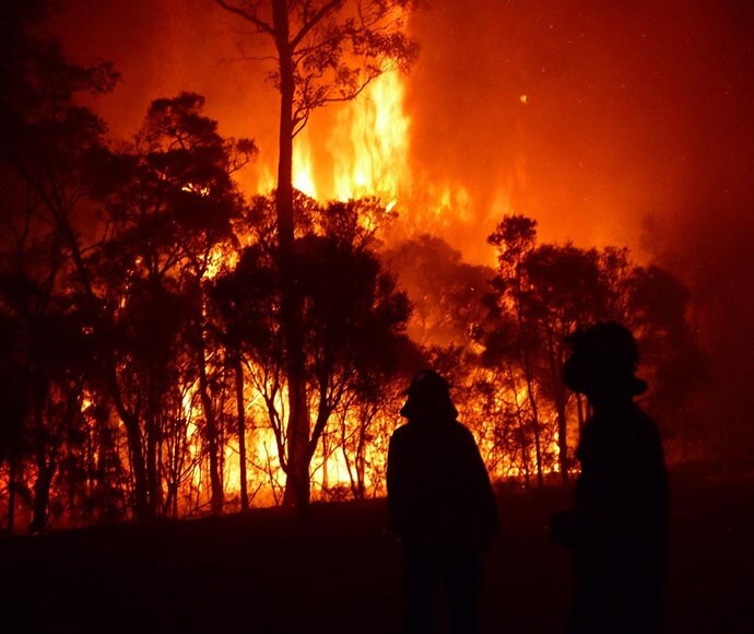 Two firemen, silhouetted against a large wall of fire at night.