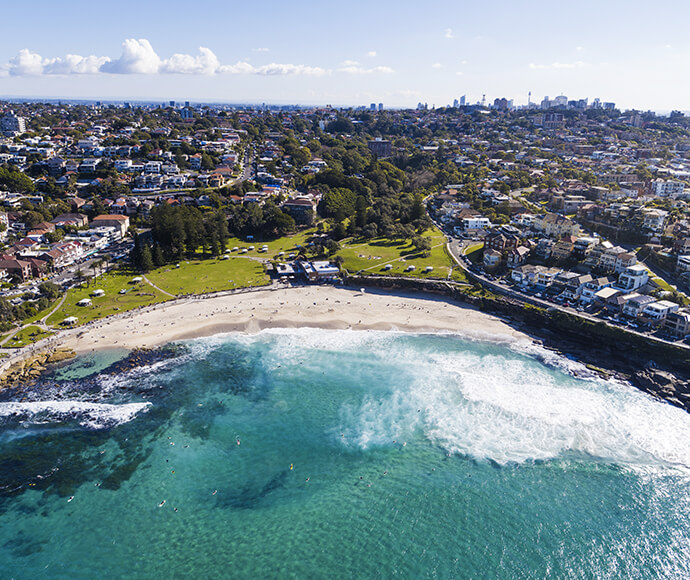 Aerial view of swimmers at the beach, you can see the large green park behind the beach and the Sydney CBD buildings far in the background