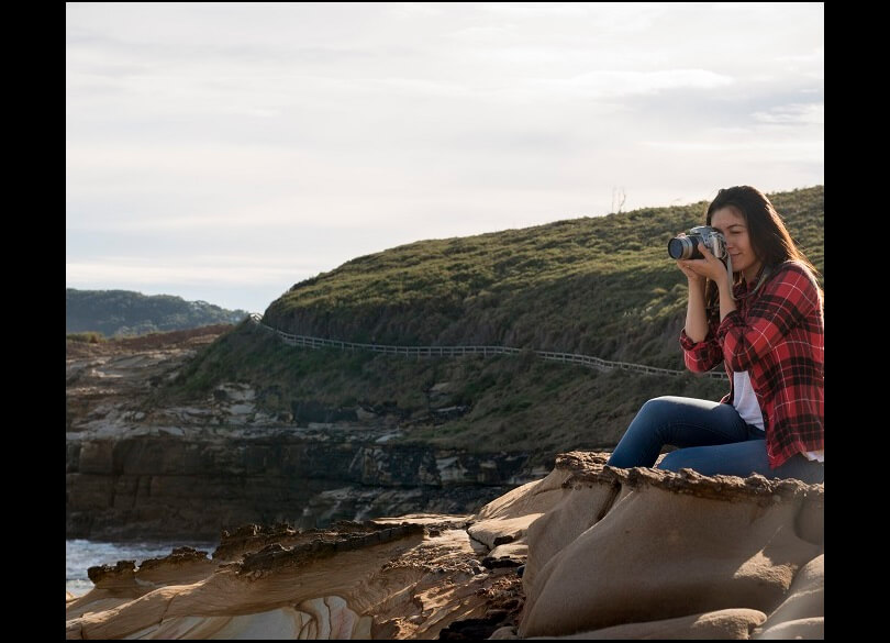 A woman sits on a rocky ledge. She takes a photo of a pretty coastal view. The view includes the Bouddi National Park coastal walk.