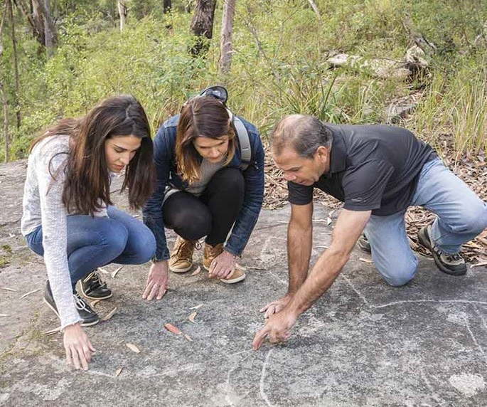 Two people are learning about the local Indigenous rock art. An Indigenous elder is examining the art with them as they crouch on the rocky ground. In the background, you can see trees and vegetation.