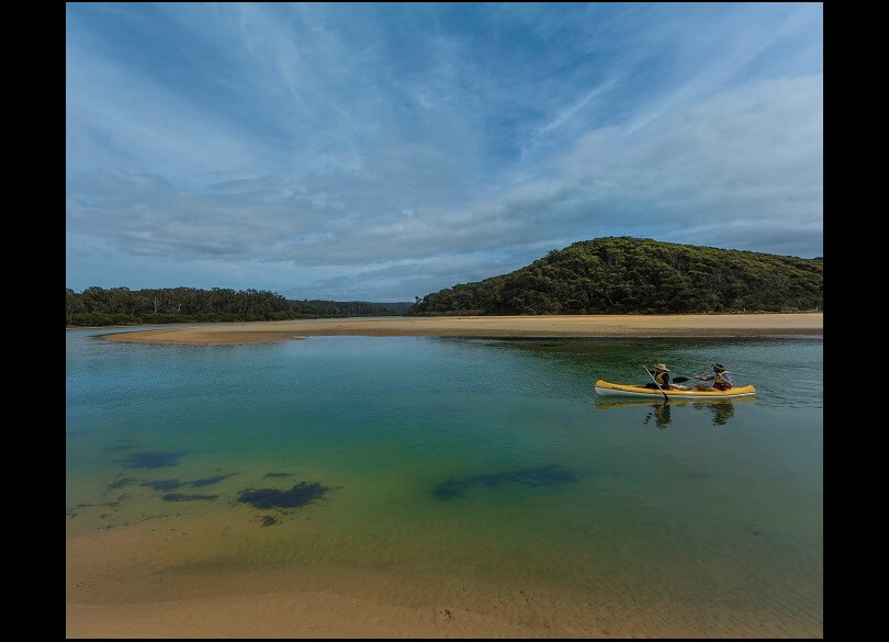 A person kayaking on a calm body of water with clear visibility to the sandy bottom, near a sandbank, with lush green hills and a blue sky with scattered clouds in the background.