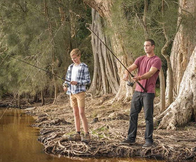 Two people fishing on the banks of a river. The background is thick forest with large trees.