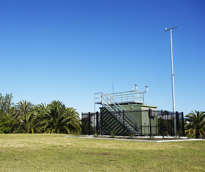 Newcastle Local Air Quality Monitoring Station, enclosed by a metal fence with a green shelter housing environmental monitoring equipment such as antennae and sensors, set amidst a grassy field with palm trees under a clear blue sky.