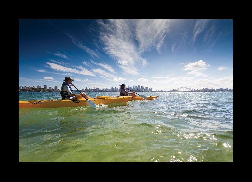 Two individuals kayaking on a clear day with a city skyline and bridge in the background, under a blue sky with wispy clouds.