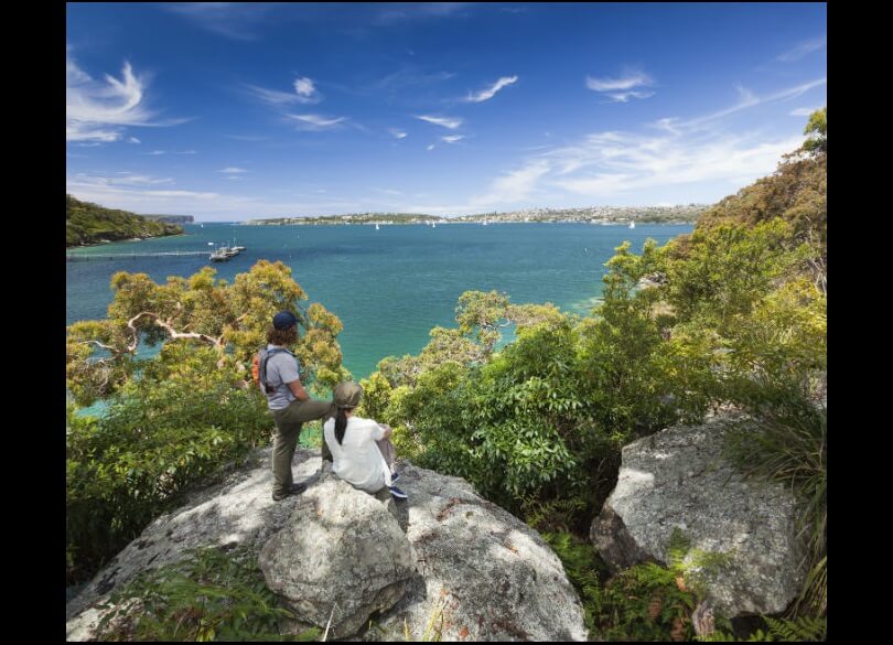 Two people enjoying the view of Sydney Harbour from the Manly scenic walk. They are sitting on a large boulder; the harbour stretches out in front of them with blue skies above.