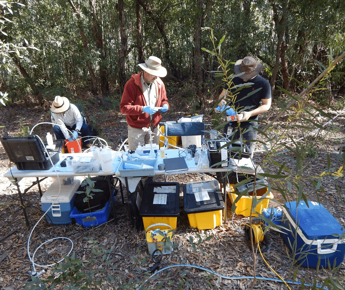 Three people with hats on outside with bush behind and scientific equipment on table infront doing sampling work.