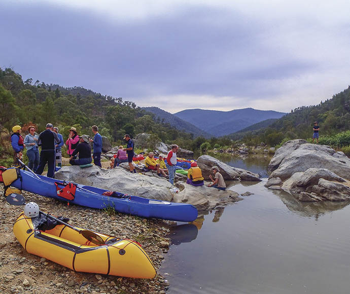 Group of people by a riverbank with two canoes on the shore, surrounded by forested mountains under an overcast sky.