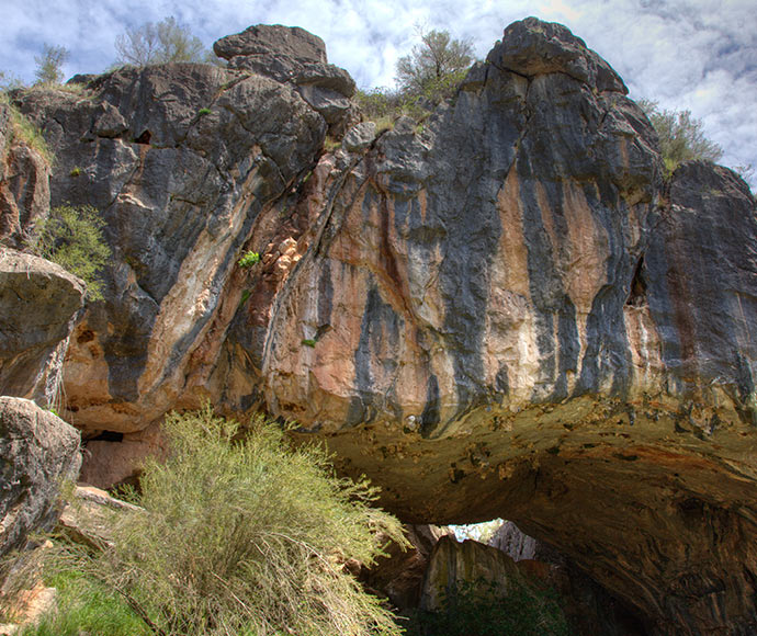 Massive limestone rock formations with vertical striations under a blue sky with clouds at Borenore Karst Conservation Reserve. A natural arch is visible at the bottom, surrounded by green vegetation.