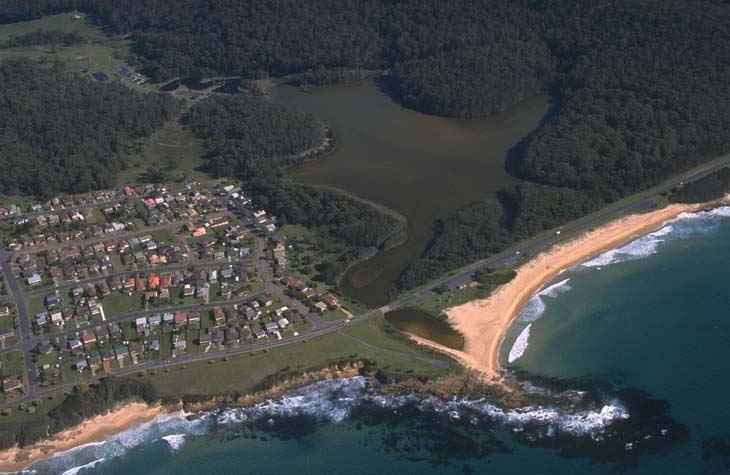 Aerial view of Kianga Lake adjacent to a sandy beach with waves, bordered by a residential area with structured streets and houses, set against a backdrop of dense green foliage.