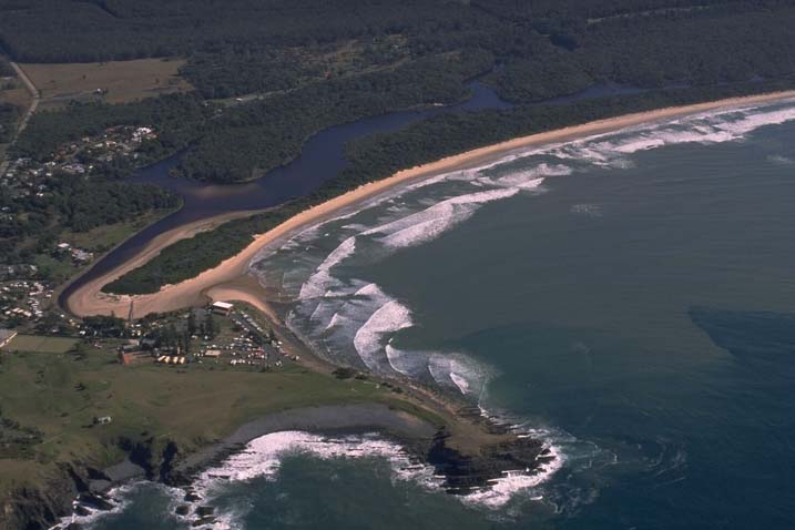 Aerial view of Killick Creek, showing its meandering path through a dense green landscape, flowing into the ocean. The creek is near a small coastal community with roads and buildings visible. The beachfront shows waves breaking gently on the shore, with a clear demarcation between the sandy beach and the lush greenery surrounding it.