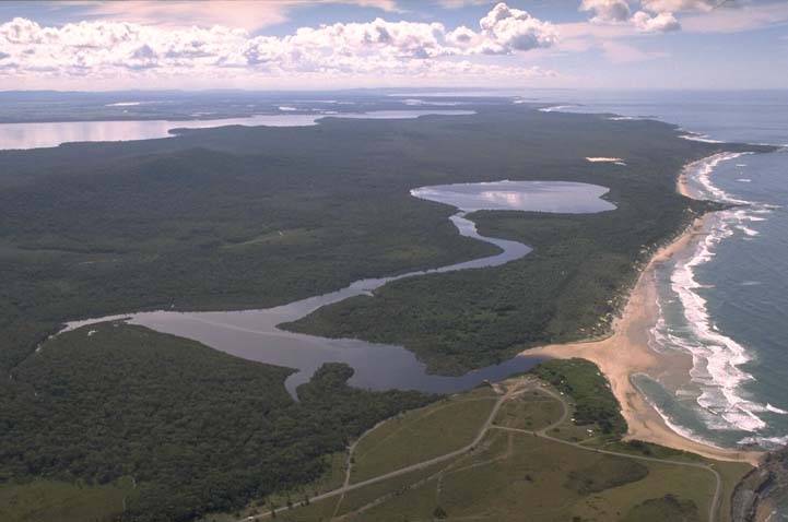 Aerial view of Lake Arragan surrounded by dense greenery, with a winding river leading to the sea. The coastline features a sandy beach with waves crashing ashore, under a sky dotted with fluffy clouds.