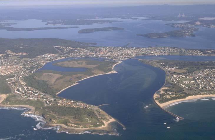 Aerial view of Lake Macquarie showing the expansive blue water body with surrounding greenery, residential areas, and peninsulas extending into the lake.