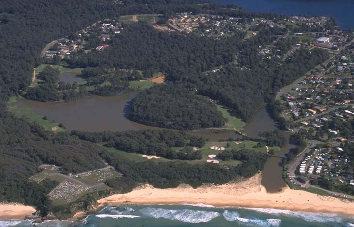 Aerial view of Little Lake in Narooma, showing a tranquil water body nestled between lush greenery with a sandy beachfront leading into the ocean. Residential areas are visible surrounding the lake, illustrating a serene coastal landscape.