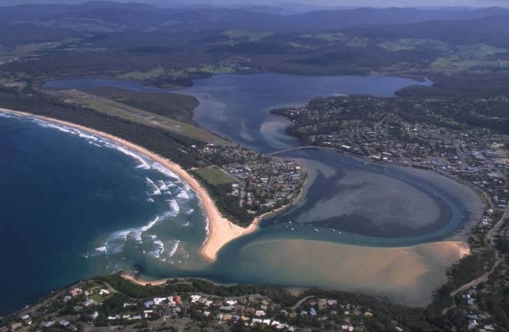 Aerial view of Merimbula Lake flowing into the ocean, with visible sandbanks at the river mouth and a nearby town along the waterways.