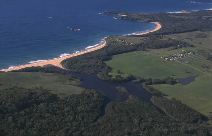 Aerial view of Meringo Creek with its winding path through green fields, leading to its mouth at a sandy beach adjacent to the clear blue ocean.