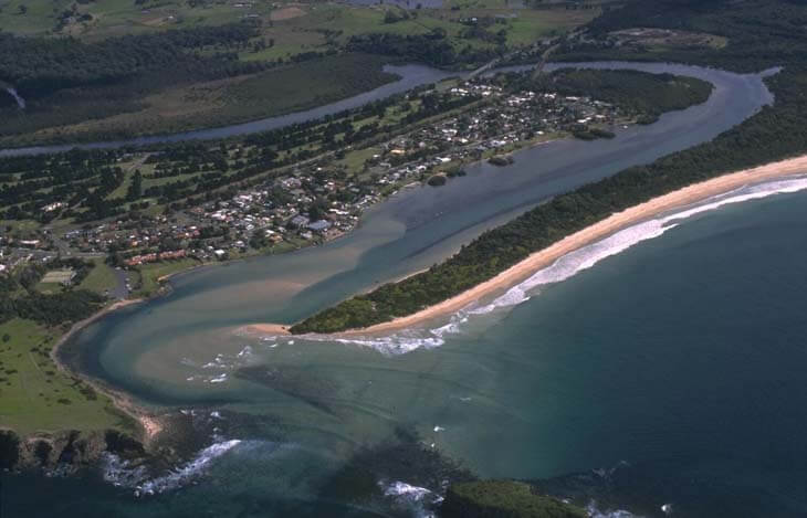 Aerial view of Minnamurra River showing its winding path through a lush landscape before meeting the ocean. The river is flanked by a small town with organized streets and houses on one side, and a sandy beach with breaking waves on the other. The contrast between the calm river waters, the structured urban area, and the dynamic coastline creates a diverse and captivating scene.