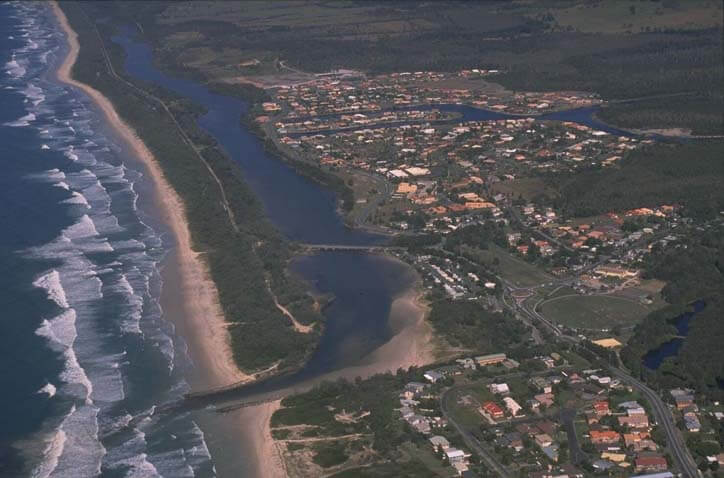 Aerial view of Mooball Creek, showing its winding path as it flows into the ocean. The creek is surrounded by a dense residential area with numerous houses and streets, and a beach running parallel to the coastline. The contrast between the natural waterway and developed land highlights the intersection of human habitation and natural landscapes.