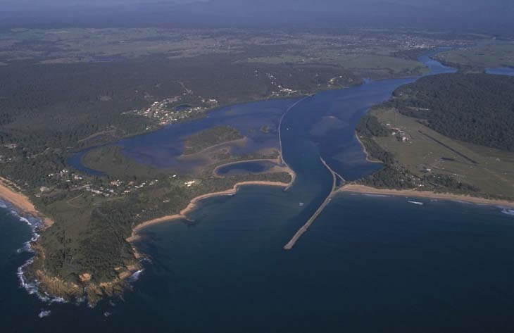 Aerial view of Moruya River, showing its winding path as it flows into the ocean. The creek is surrounded by a residential area with numerous houses and streets, and a beach running parallel to the coastline.