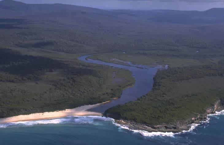 An aerial view of Nadgee River, near the coast. A dense forest is in the background. A sandy beach separates the calm lake from the wavy ocean.