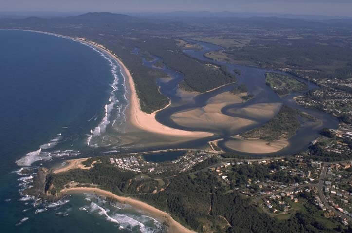 Aerial view of Nambucca River, showing a beachfront with waves crashing onto the shore, a river meandering through the landscape, residential areas with numerous houses, greenery, and roads.