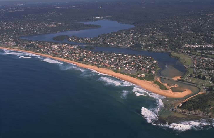 Aerial view of Narrbeen Lagoon, showing a beachfront with waves crashing onto the shore, a river meandering through the landscape, residential areas with numerous houses, greenery, and roads, and a clear boundary where the urban area meets the natural water bodies.