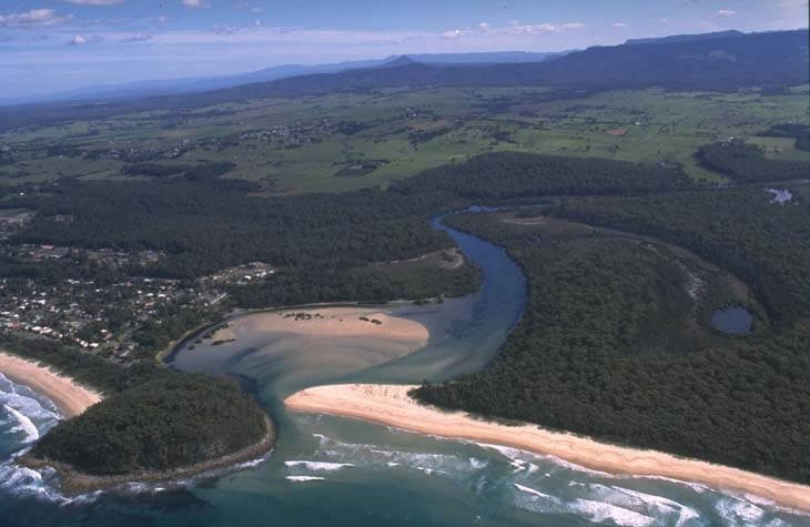 Aerial view of Narrawallee Inlet, showing a beachfront with waves crashing onto the shore, a river meandering through the landscape, residential areas with numerous houses, greenery, and roads, and a clear boundary where the urban area meets the natural water bodies.