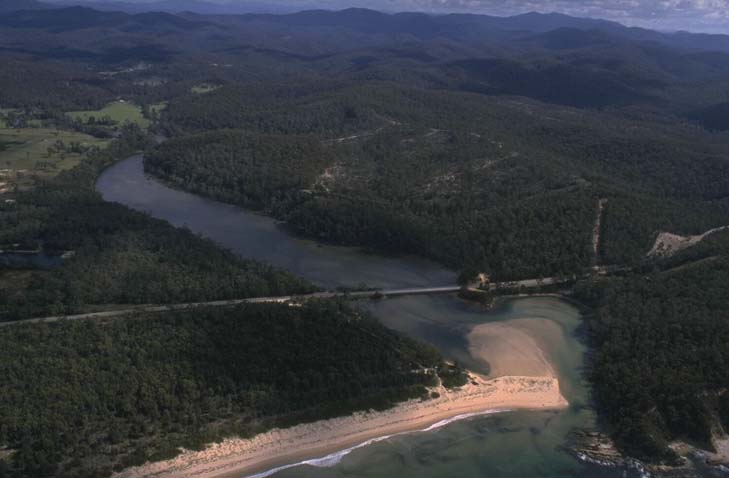 Aerial view of the Nullica River winding through a dense forest with mountains in the background. A bridge crosses over a section of the river, and a sandy beach area is visible at the river’s bend.