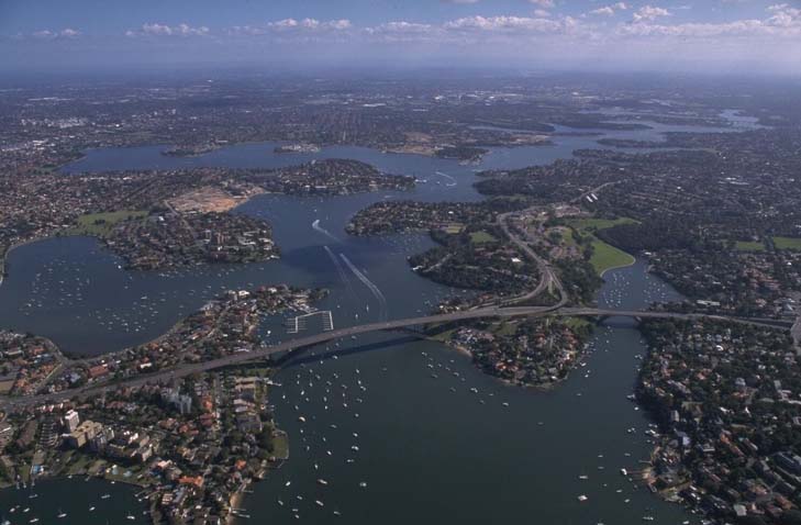 Aerial view of Parramatta River in Australia, showcasing a wide river meandering through a densely populated landscape. The river is flanked by numerous boats and waterfront properties, with bridges connecting the two sides. The surrounding area is a mix of green spaces and urban development, with the skyline fading into the horizon under a clear blue sky.