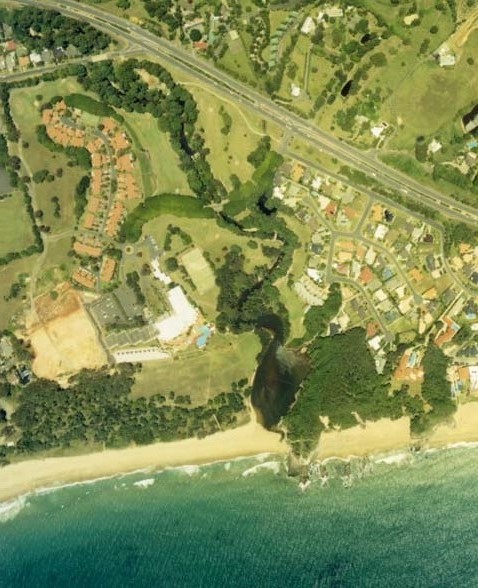 Aerial view of Pine Brush Creek near a coastal area, showcasing a contrast between the natural waterway, greenery, and the adjacent developed land with buildings and roads.