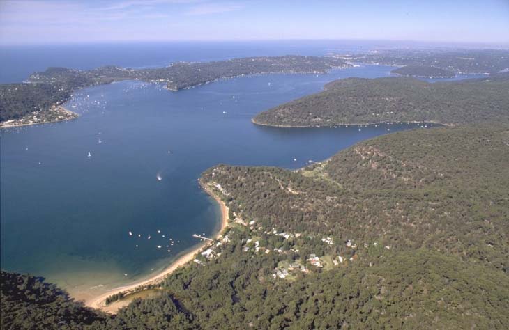 Aerial view of Pittwater, a coastal estuary region north of Sydney, Australia, featuring a vast blue water body surrounded by lush greenery with boats scattered across the water and a small beach visible along the shoreline.