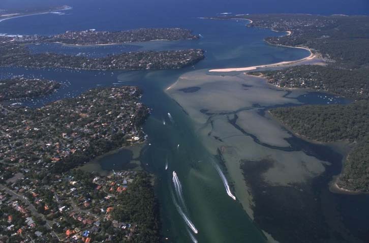 Aerial view of Port Hacking, showing the winding waterways with boats leaving white trails, surrounded by dense greenery and residential areas.