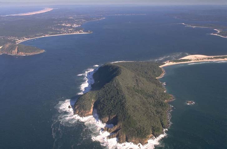 Aerial view of Port Stephens, showcasing a large natural harbor with a narrow entrance bordered by land with dense vegetation. The water is a deep blue with visible waves crashing against the rocky shoreline. Sandy beaches are visible in the distance, and the overall landscape suggests a serene coastal ecosystem.