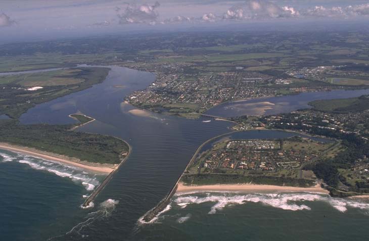 Aerial view of Richmond River meeting the sea, showcasing the contrast between the brown river waters and blue ocean. The landscape includes patches of greenery and urban development, highlighting the interaction between natural and human-made environments.