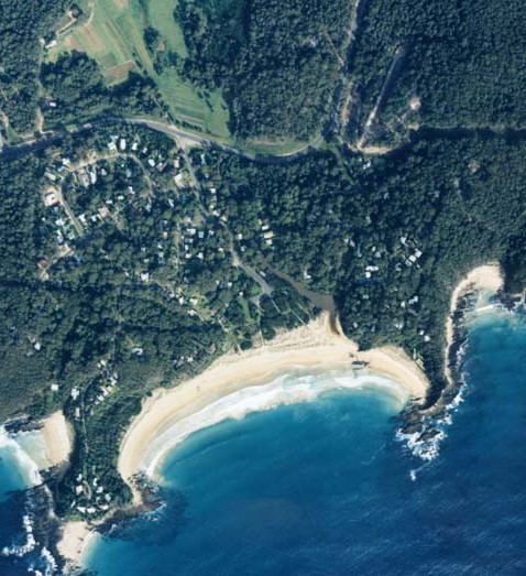 Aerial view of Saltwater Creek in Rosedale showing a curved sandy beach bordered by dense greenery and trees, with clear blue waters on one side and a cluster of houses amidst the foliage on the other.