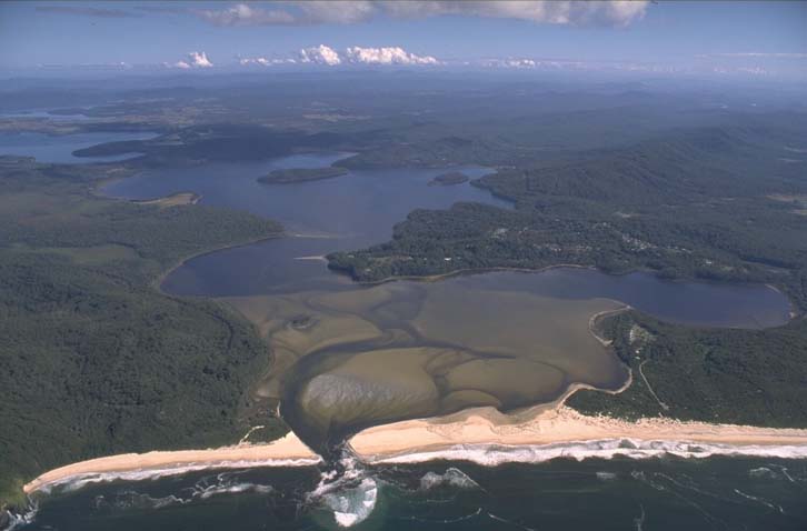 Aerial view of Smiths Lake, a coastal lagoon bordered by a sandy beach with the ocean on one side and dense green forest surrounding the water. The lake’s surface reflects the sky, and there are visible sandbars within it. In the distance, clouds hover over the horizon.