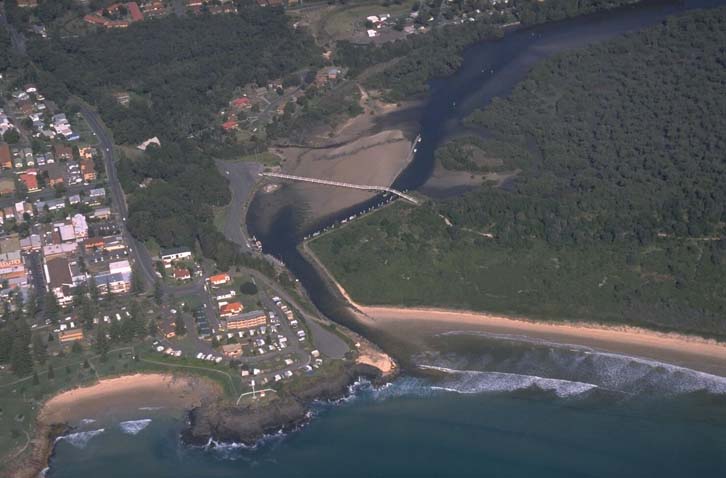 Aerial view of South West Rocks Creek, showing the convergence of a winding creek with the ocean. A bridge spans the narrow point of the creek, surrounded by dense greenery on one side and a sandy beach on the other. Residential areas with several buildings are visible near the coastline, contrasting natural and developed land.