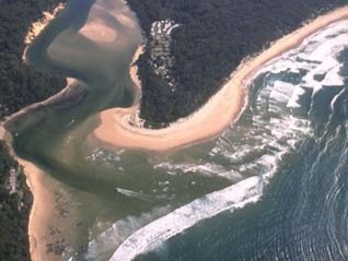 Aerial view of St Georges Basin showing a coastal water body with a narrow inlet leading to the open sea. The basin is surrounded by dense greenery and forested areas, with sandy shores visible at the bottom of the image where the basin meets the sea. Waves can be seen breaking on the shoreline, indicating an active coastal environment. The topography suggests a natural and possibly protected area, with no immediate signs of urban development in this section of the landscape.