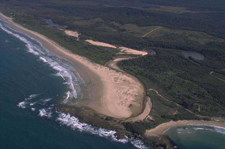 Aerial view of Station Creek showing a wide sandy beach with gentle waves breaking along the shoreline. The creek meanders through a dense green forest before emptying into the ocean. The contrast between the lush forest, sandy beach, and clear blue water creates a striking natural landscape.