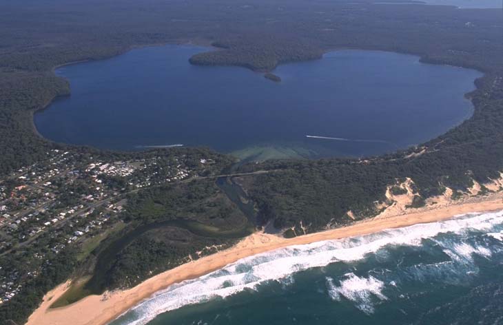 Aerial view of a large, circular blue lake surrounded by dense green forests with a small town on one side. A narrow strip of sandy beach separates the lake from the ocean, with waves visible along the shoreline.