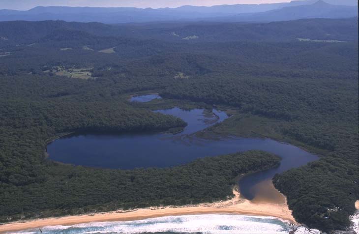 Aerial view of Termeil Lake, featuring a large dark blue lake surrounded by dense green forest. A narrow river connects the lake to a beach with light brown sand at the bottom of the image. The beach meets the ocean, and waves are visible. In the distance, there are rolling hills with a hazy blue appearance under a clear sky.