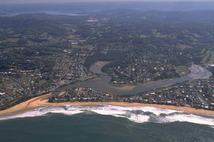 Aerial view of Terrigal Lagoon with a winding river leading into it, surrounded by dense greenery and residential areas. The lagoon is near a beachfront with breaking waves, highlighting the diverse landscape of coastal and inland water bodies juxtaposed with urban development.