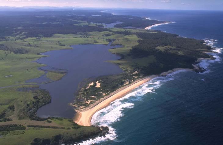 Aerial view of Tilba Tilba Lake estuary showing a tranquil body of water with branching inlets surrounded by lush greenery. A narrow strip of sandy beach separates the lake from the open sea, with waves crashing onto the shore. The horizon reveals a clear sky above and hints at a vast ocean extending beyond.