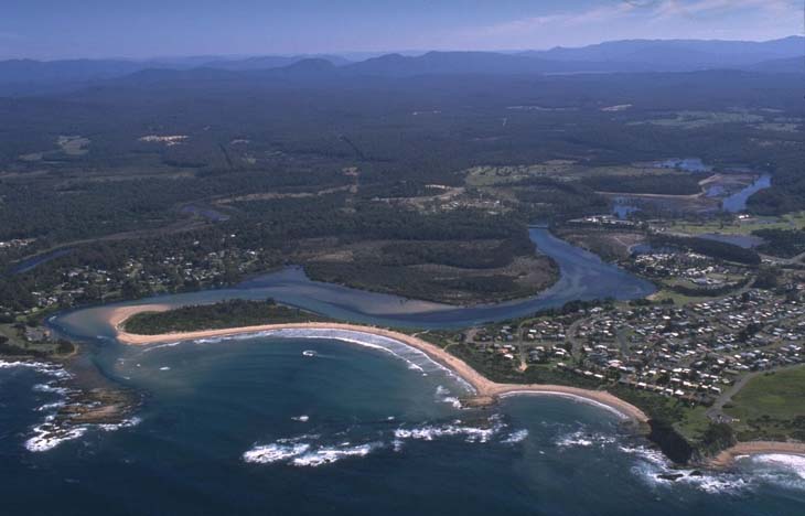 An aerial view of the Tomaga River estuary, where the winding river meets a larger body of water, likely the ocean. The estuary is surrounded by sandy beaches forming a curved peninsula that partially encloses the river’s mouth. Dense greenery, possibly forests or bushland, is visible, along with a residential area featuring numerous buildings.