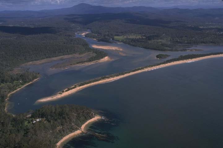 Aerial view of the Towamba River winding through dense greenery and forested areas, flowing into a larger body of water. The riverbanks are sandy, and small structures or buildings are visible near the shoreline. Rolling hills or low mountains are seen in the distance under a partly cloudy sky.