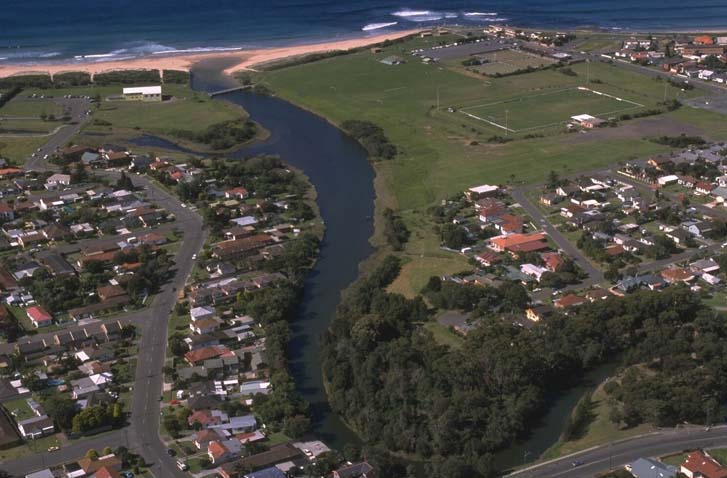 Aerial view of Towradgi Creek meandering through a landscape with residential areas on one side and a beachfront on the other. The creek flows into the ocean, with green spaces including parks or fields adjacent to the residential area.