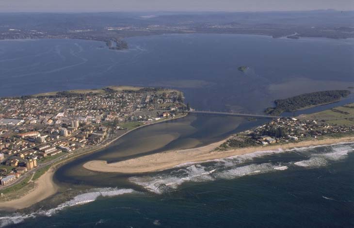 Aerial view of Tuggerah Lake, a large coastal lagoon on the Central Coast near Sydney, New South Wales. The lake is connected to the Tasman Sea by a short channel. In the foreground, there is a sandy beach adjacent to an urban area with buildings and streets. The lake is expansive, with land visible on the far side and a peninsula extending into it.